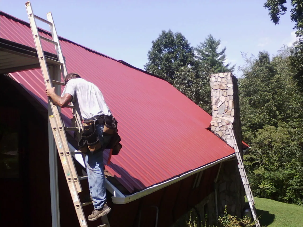 Roofer on a ladder beside a red metal roof and stone chimney — Soffit & Fascia Repair by Roofing Brothers in Wheat Ridge